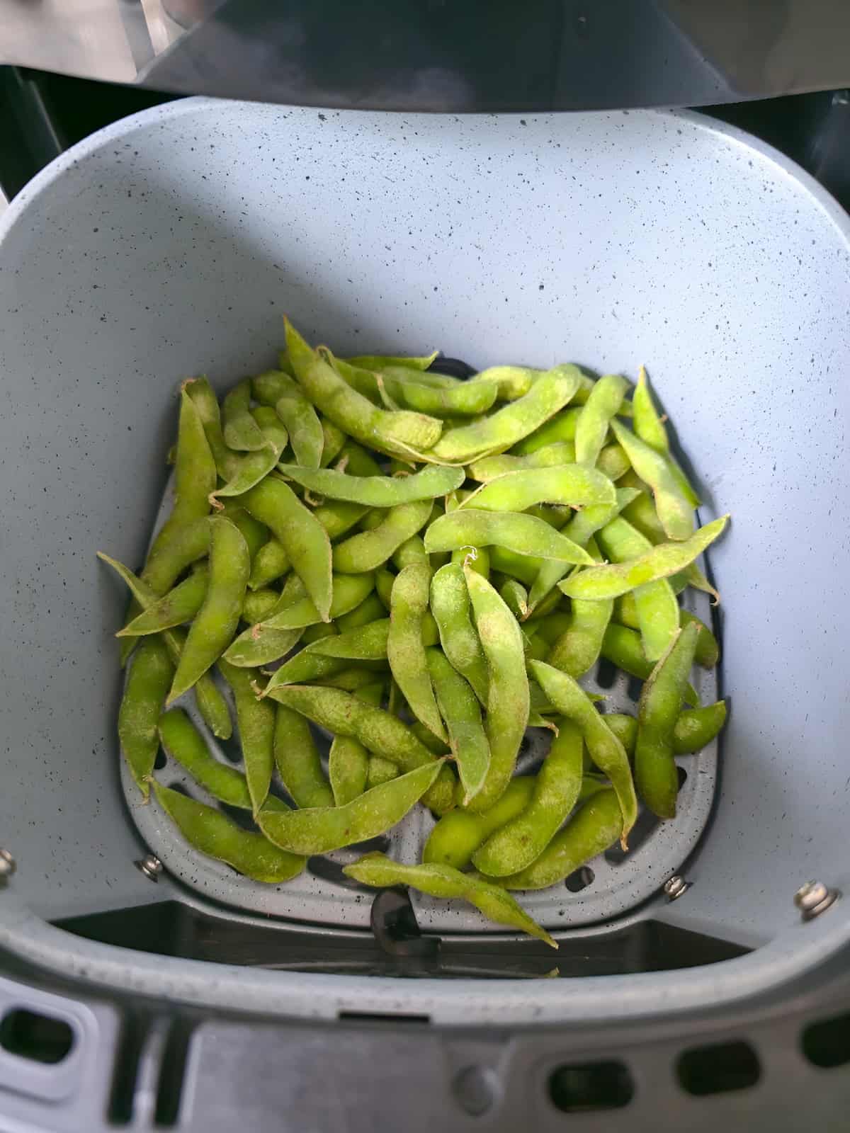 Frozen edamame spread in an air fryer basket before cooking.