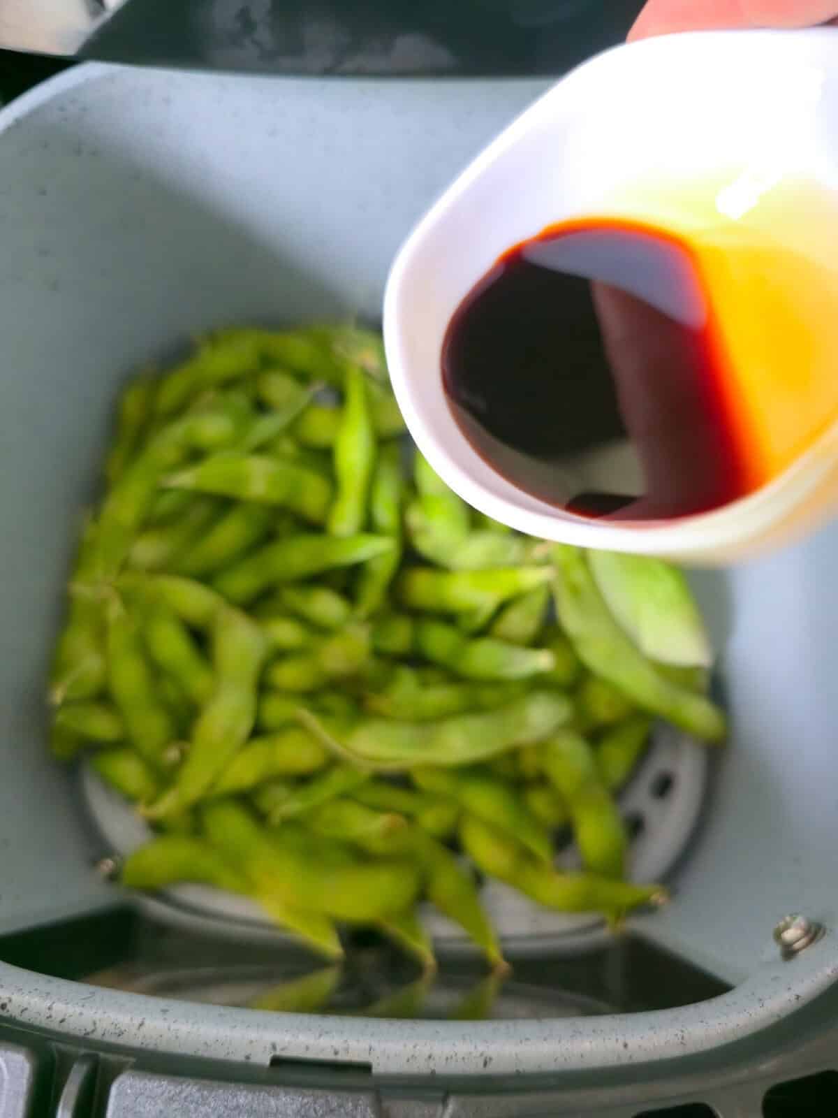 Soy sauce being poured over edamame in an air fryer basket.