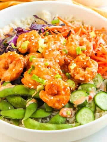 Bang bang shrimp bowl with rice, crispy shrimp, edamame, cucumbers, carrots, and purple cabbage, drizzled with creamy sauce.
