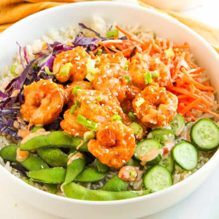 Bang bang shrimp bowl with rice, crispy shrimp, edamame, cucumbers, carrots, and purple cabbage, drizzled with creamy sauce.
