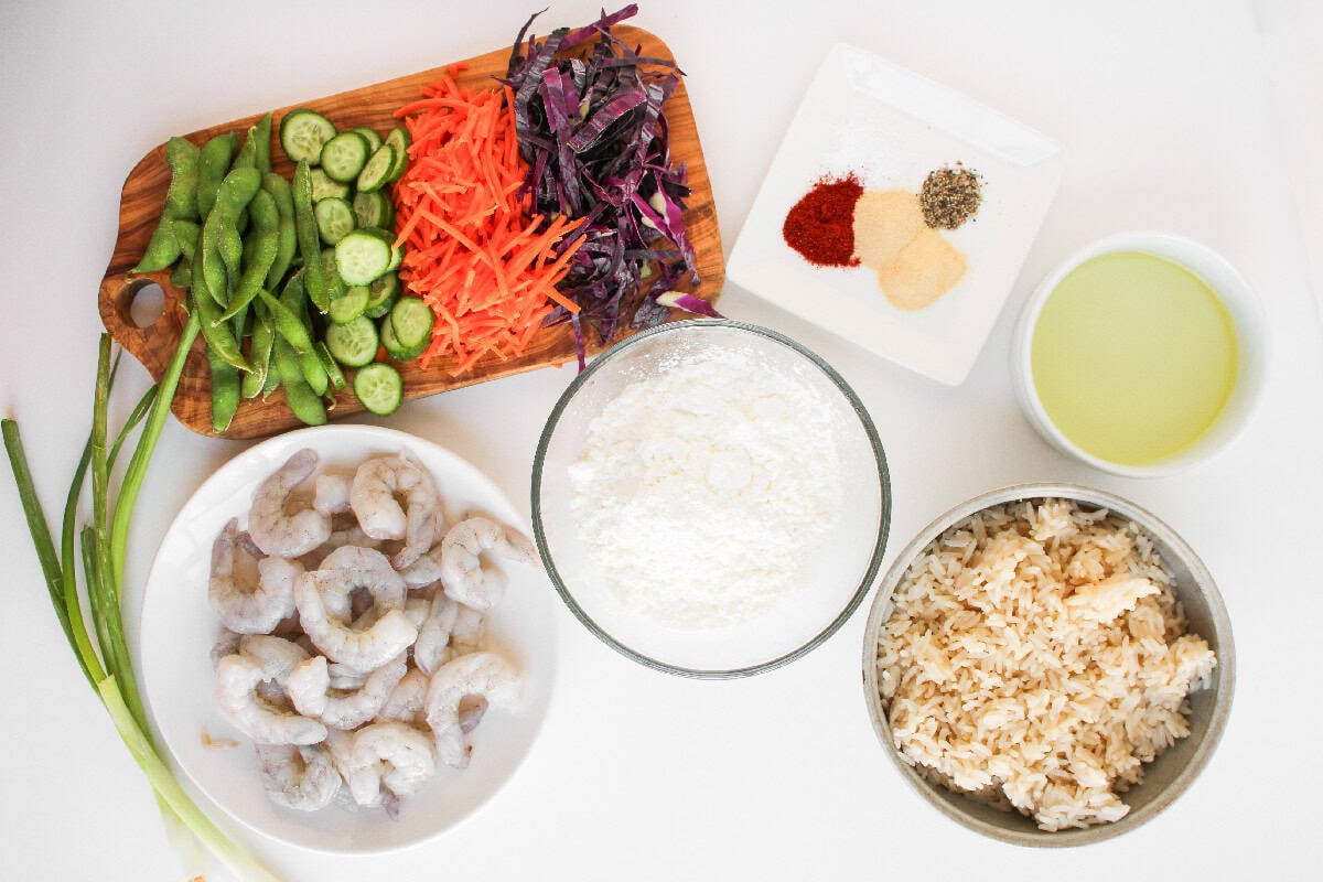 Bang bang shrimp bowl ingredients including raw shrimp, vegetables, rice, cornstarch, oil, and seasonings arranged on a white surface.