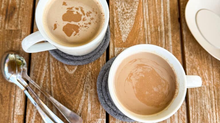 Two mugs of hot cocoa coffee served on coasters with spoons on a wooden table.