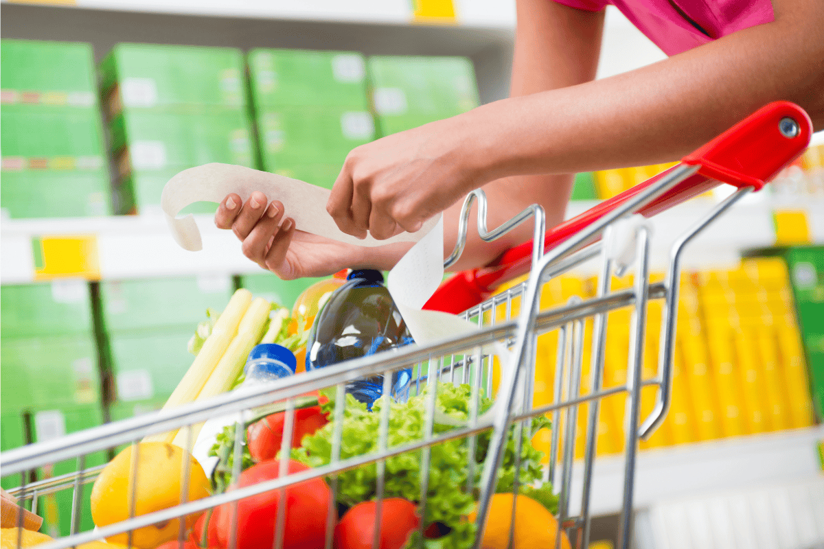 Person holding a shopping receipt over a grocery cart filled with fresh produce and bottled drinks in a supermarket.