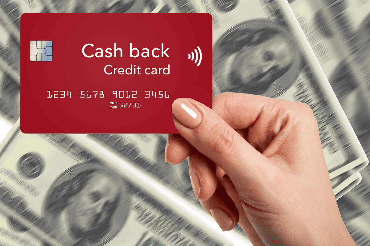 Hand holding a red cashback credit card in front of blurred U.S. dollar bills.