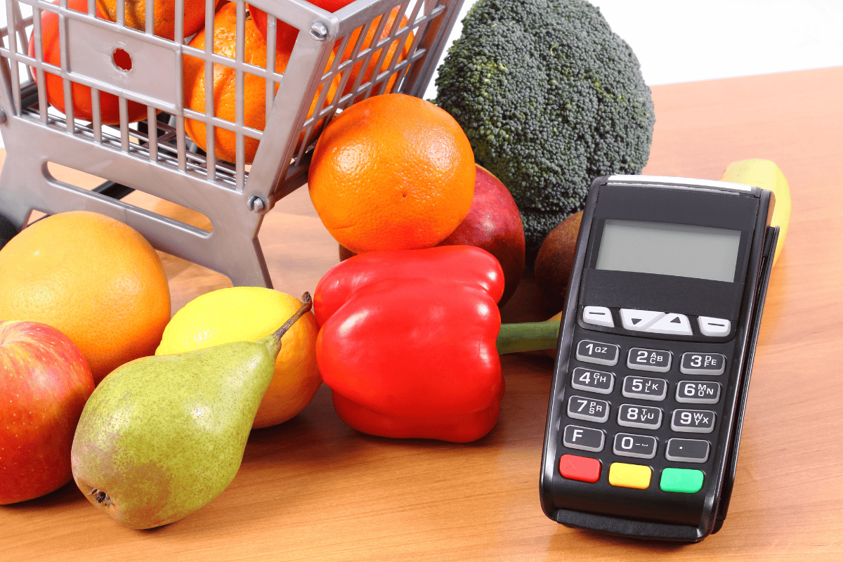Grocery cart with fresh fruits and vegetables beside a card payment terminal on a table.