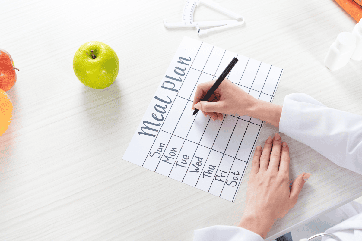 Person writing a weekly meal plan on a planner beside fresh fruit on a table.