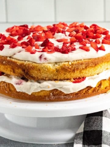 Strawberry banana layer cake with vanilla sponge, whipped cream frosting, and fresh chopped strawberries on top, displayed on a white cake stand.