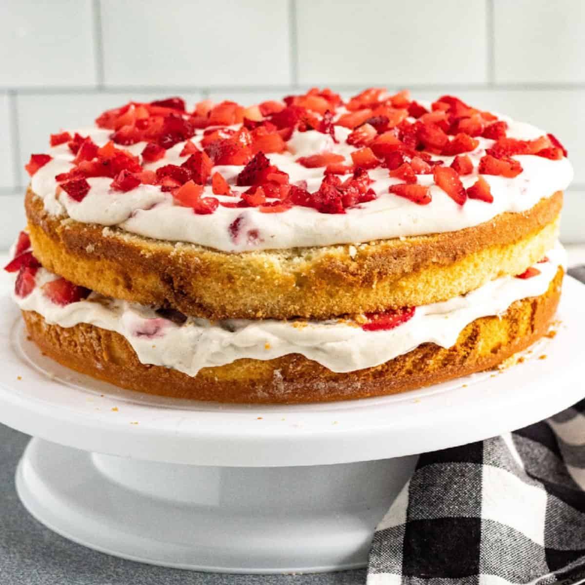 Strawberry banana layer cake with vanilla sponge, whipped cream frosting, and fresh chopped strawberries on top, displayed on a white cake stand.