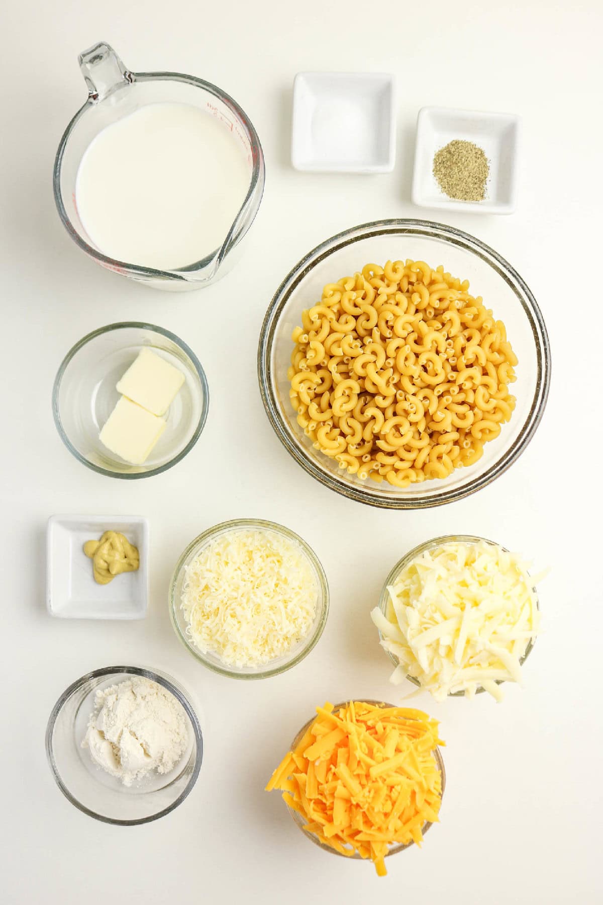 Top-down view of uncooked elbow macaroni, milk, butter, flour, mustard, salt, pepper, and three cheeses arranged on a white background.