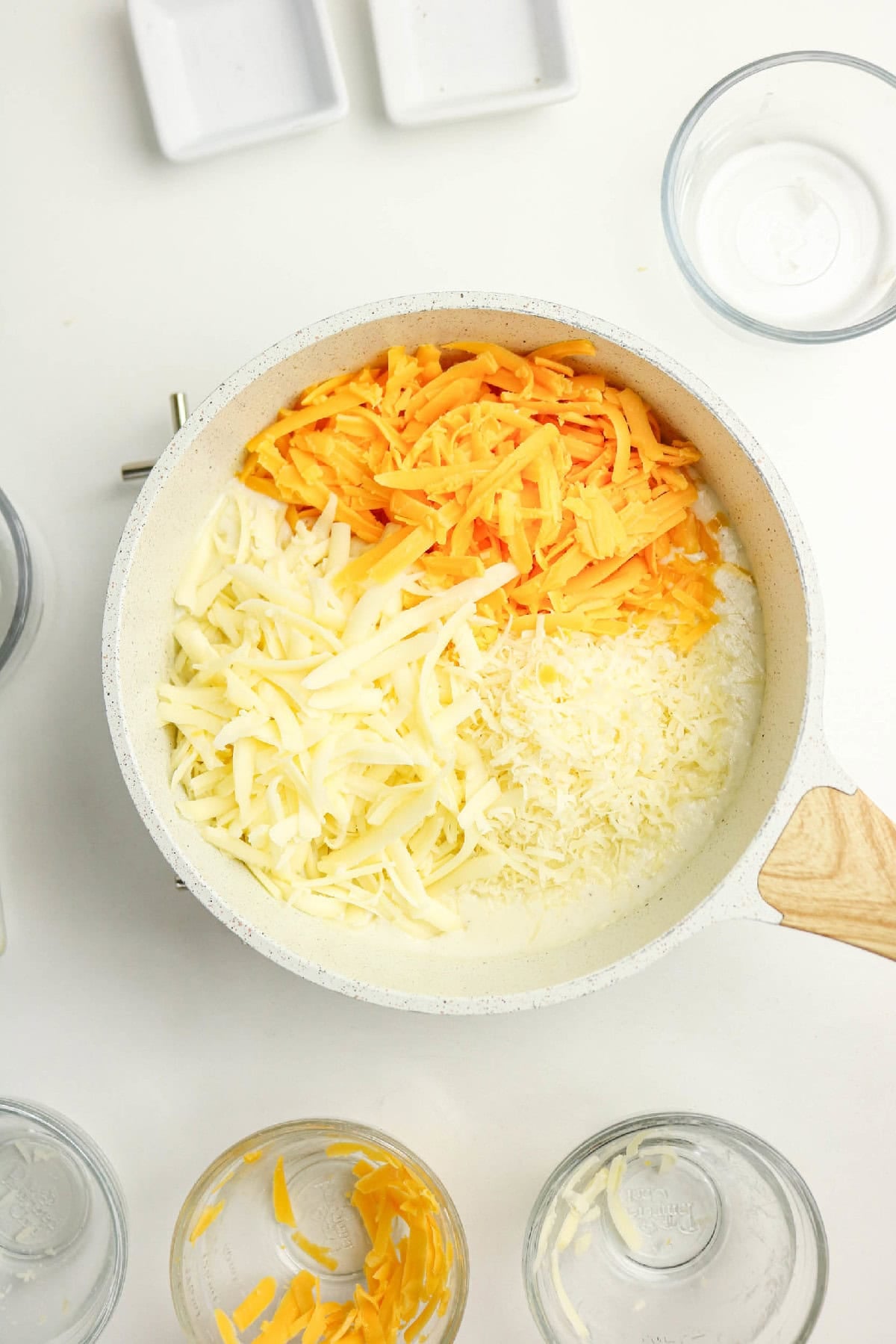 Shredded cheddar, mozzarella, and Parmesan added to the creamy sauce in a saucepan.