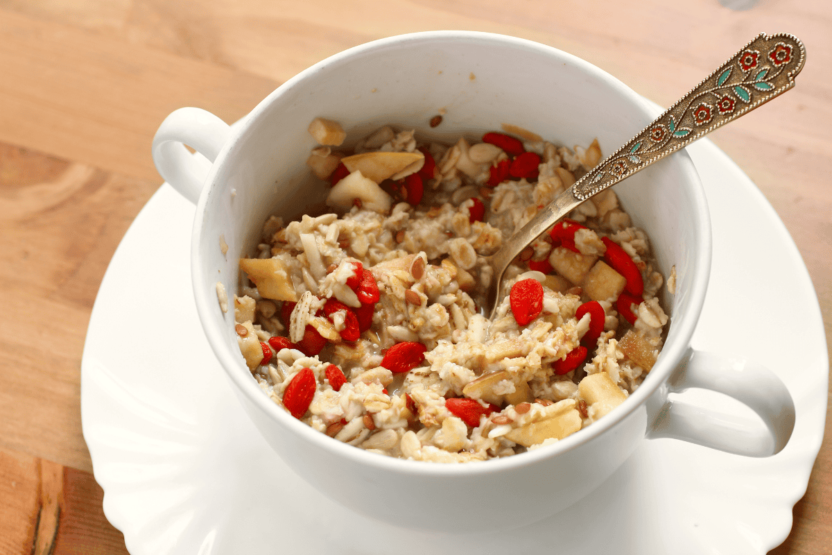 A cup of oatmeal topped with apple pieces, nuts, and dried berries sits on a white saucer with a spoon.