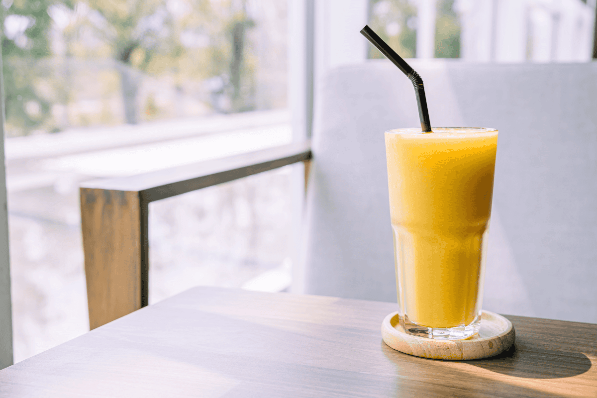 A glass of fresh orange smoothie with a straw sits on a wooden table, illuminated by sunlight.