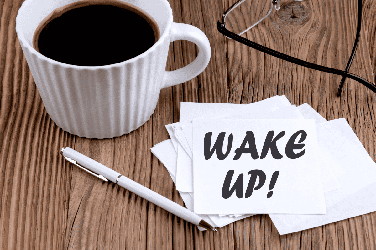 A cup of black coffee on a wooden table beside a pen, eyeglasses, and stacked cards reading “Wake Up!”