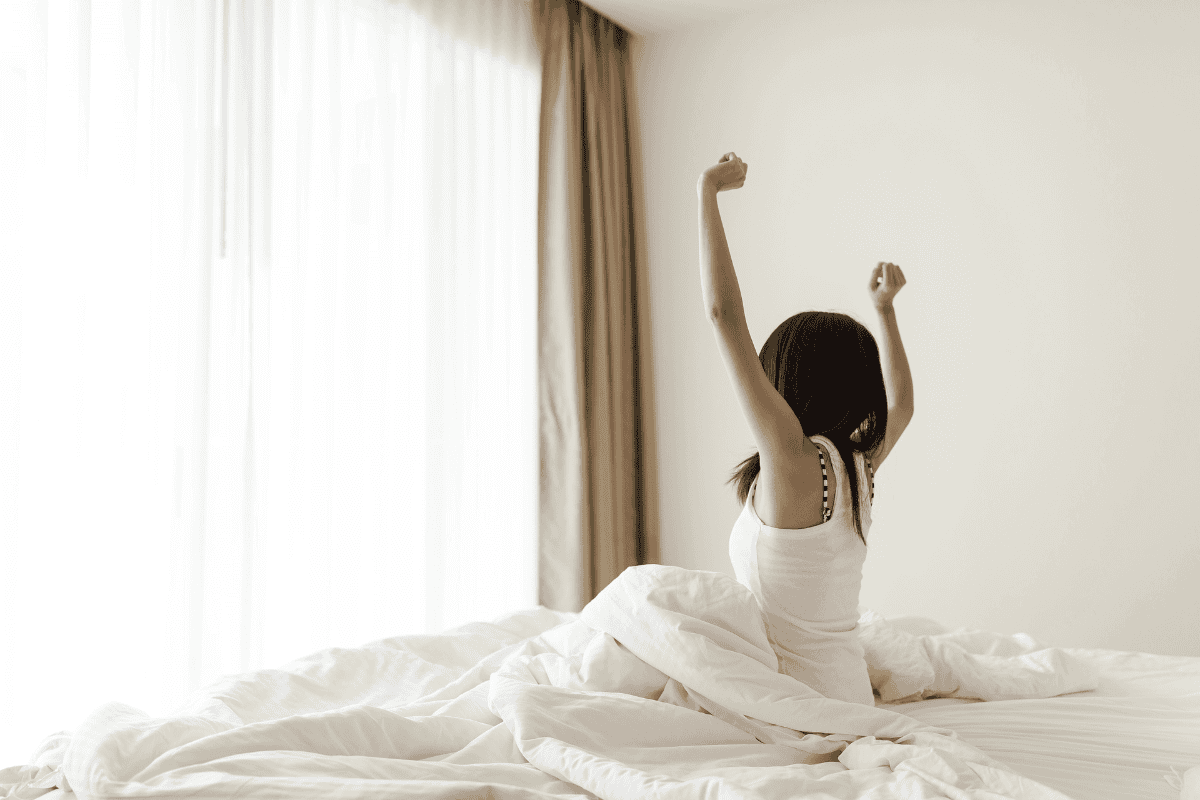 A woman stretches her arms while sitting up in bed as soft morning light fills the bedroom.