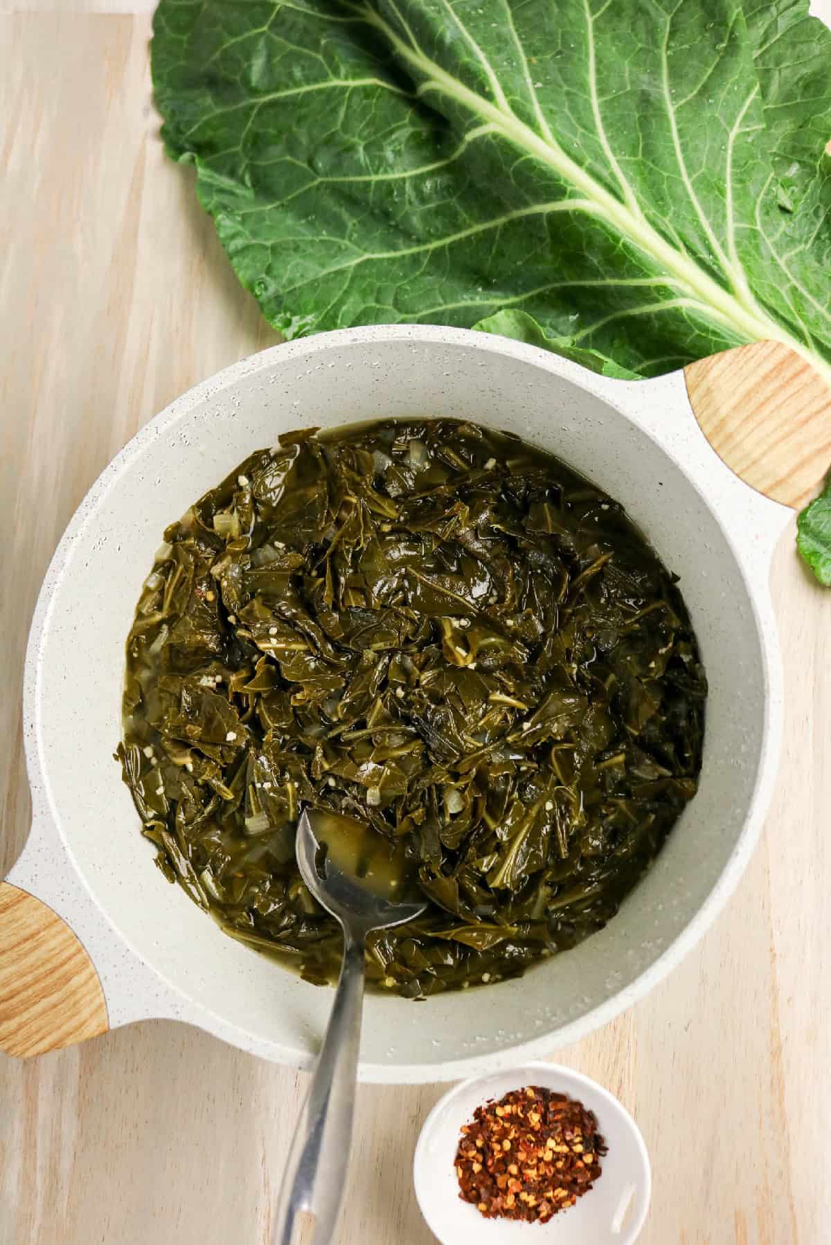 Braised collard greens cooking in a saucepan with fresh collard leaves nearby.
