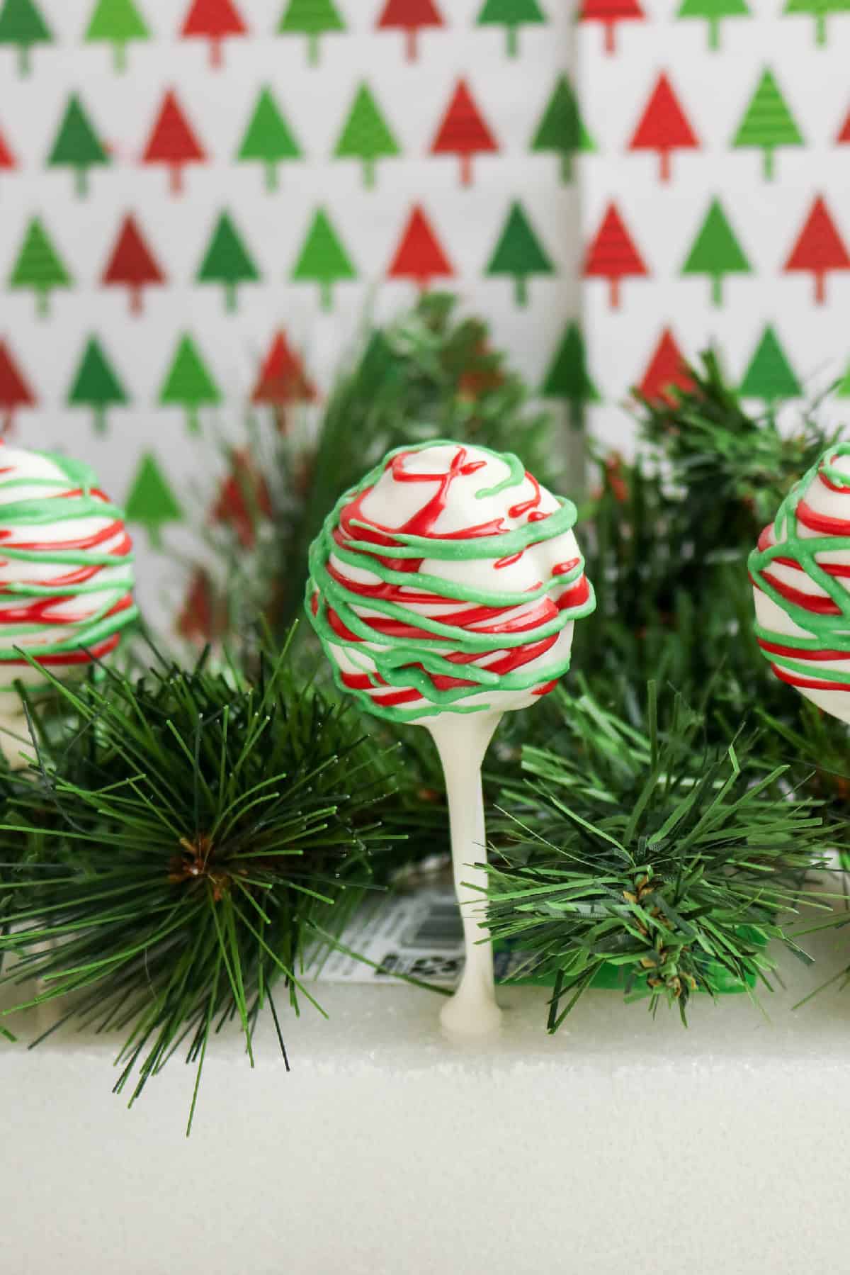 Holiday cake pops displayed on sticks with red and green candy drizzle and Christmas tree backdrop.