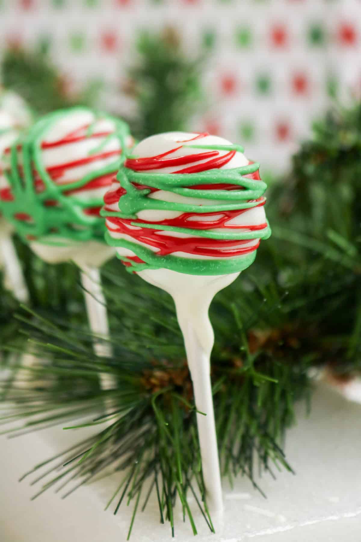 Festive cake pop with red and green drizzle on a stick, surrounded by holiday greenery.