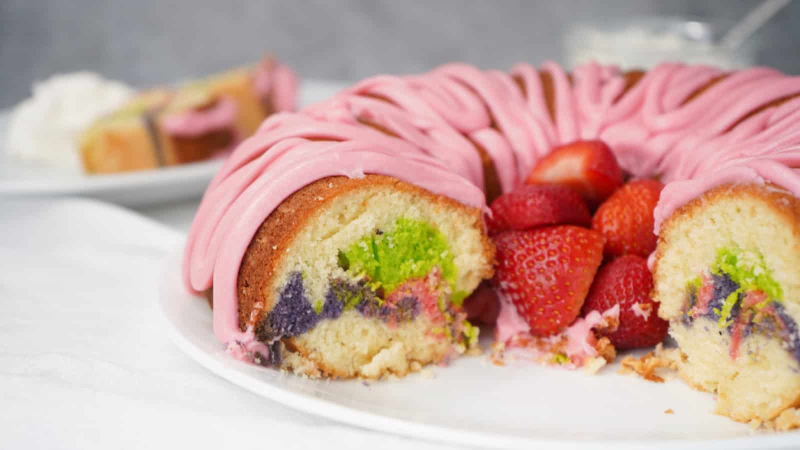 Easter bundt cake with pink icing and colorful marble swirl, sliced and served with fresh strawberries on a white plate.