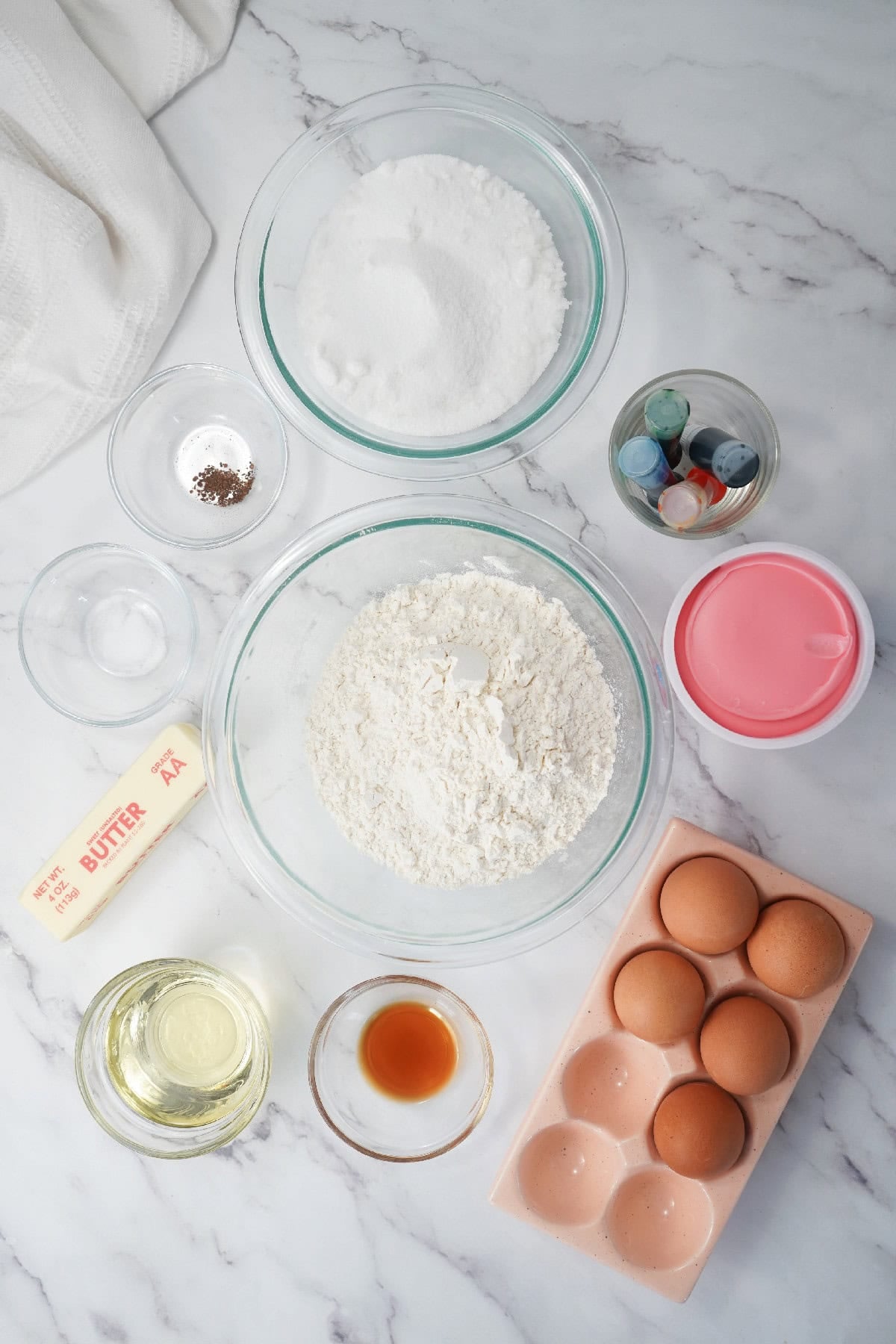 Ingredients for Easter bundt cake including softened butter, flour, eggs, and oil in glass bowls on a marble surface.