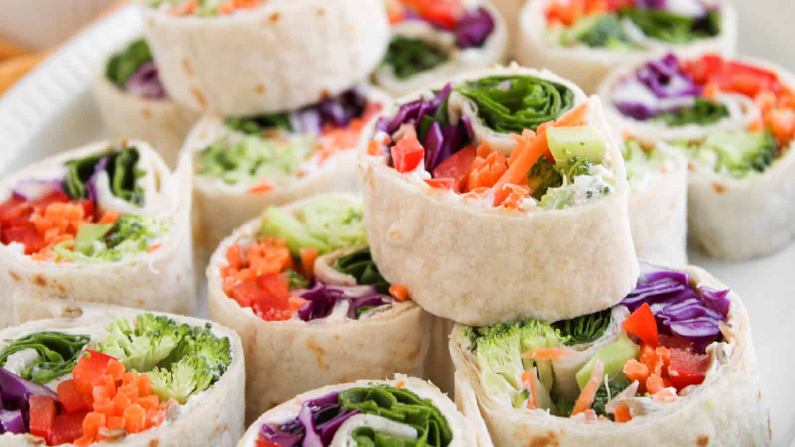Colorful vegetable wrap pinwheels with broccoli, carrots, red cabbage, and spinach stacked on a white plate.