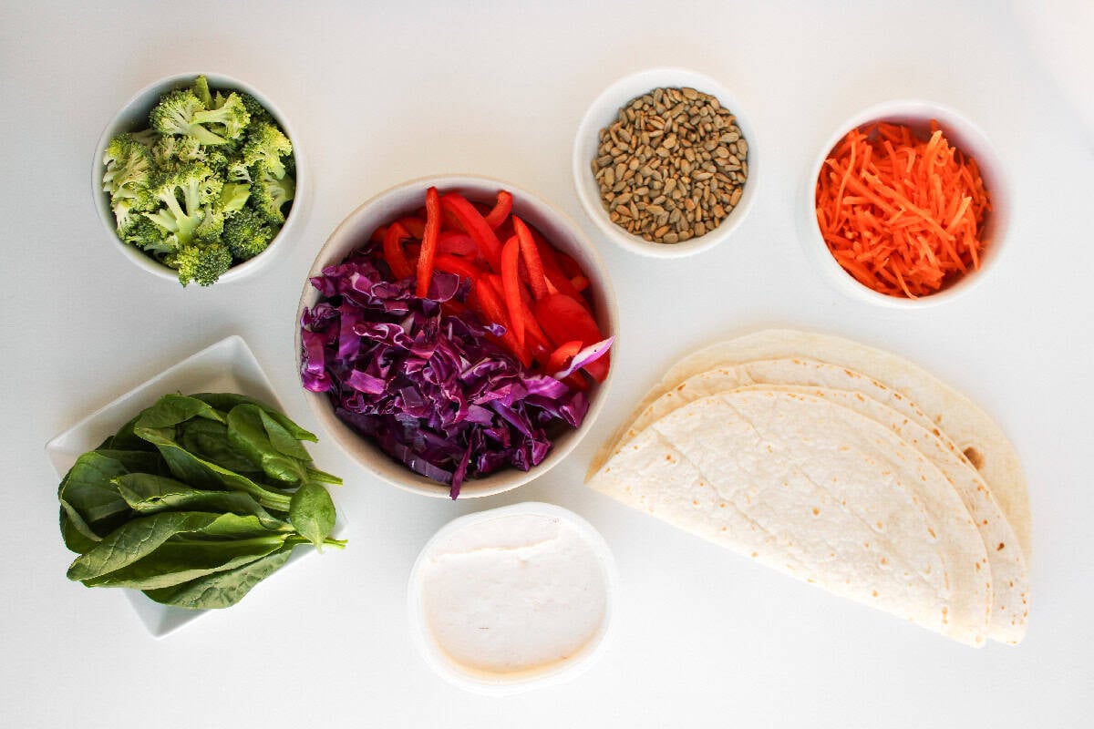 Ingredients for veggie pinwheels including flour tortillas, creamy spread, broccoli, spinach, shredded carrots, red cabbage, red bell peppers, and sunflower seeds arranged on a white background.