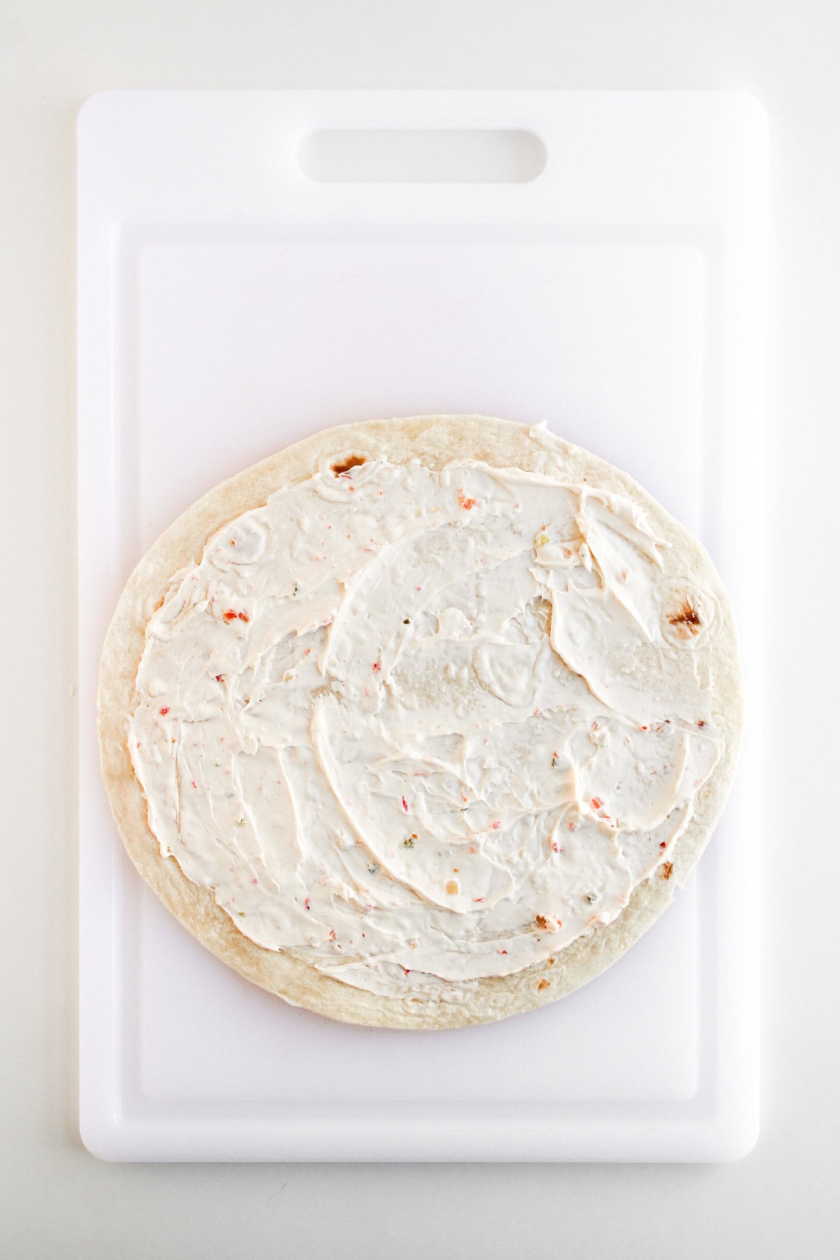 Flour tortilla evenly coated with creamy vegetable filling on a white cutting board.