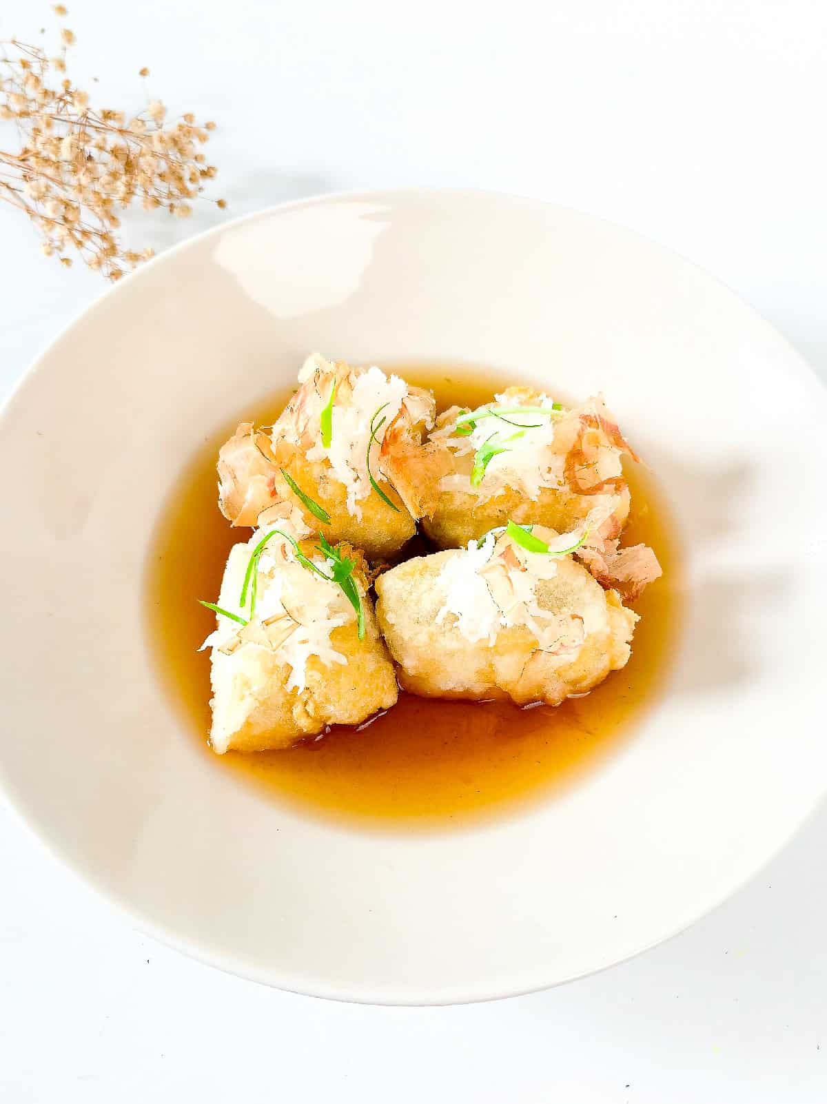 Four pieces of agedashi tofu in a shallow bowl with savory dashi sauce and toppings.