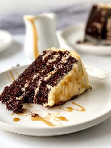 Slice of chocolate cake layered with creamy filling on a white plate, drizzled with caramel sauce, with additional slices blurred in the background.