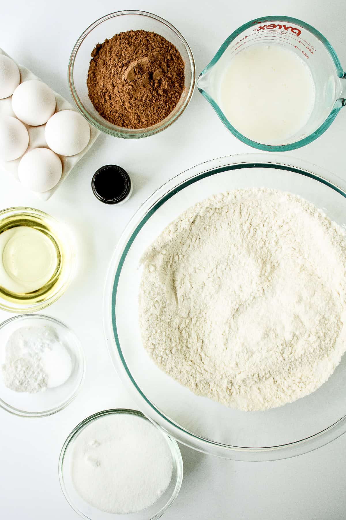Overhead view of chocolate cake ingredients arranged on a white surface, including a large bowl of flour, cocoa powder, milk in a measuring cup, eggs, oil, vanilla extract, sugar, and baking powder in small glass bowls.