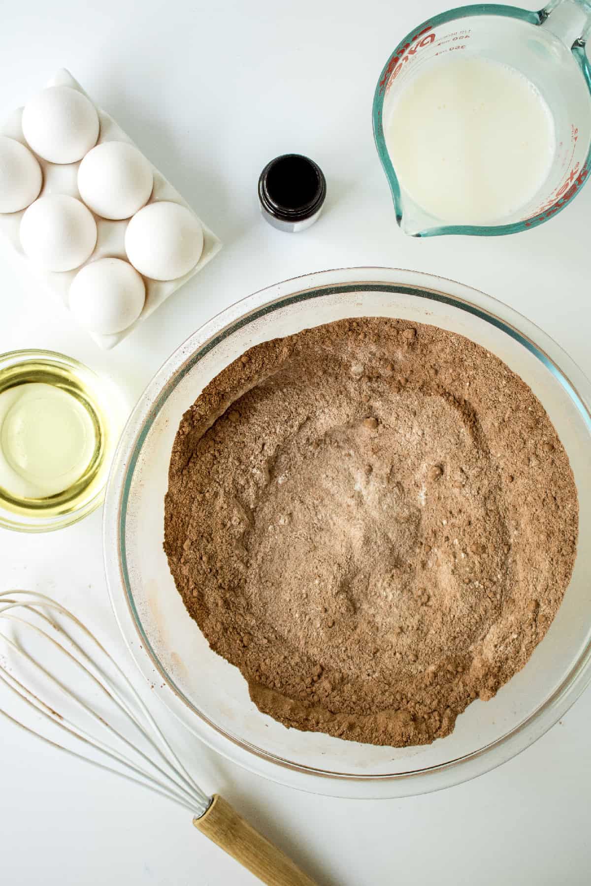 Dry chocolate cake ingredients whisked together in a large glass mixing bowl on a white surface.