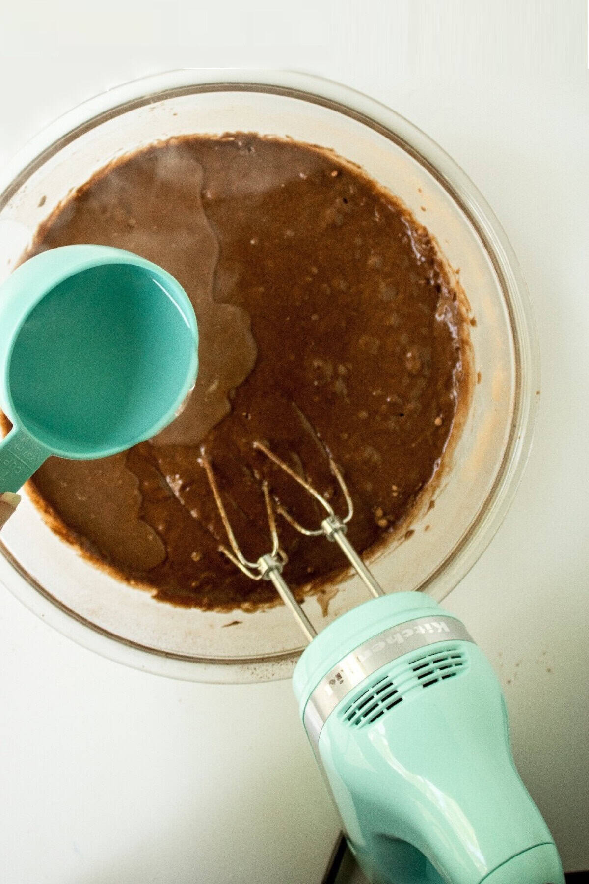 Water being poured into chocolate cake batter while mixing with an electric hand mixer.