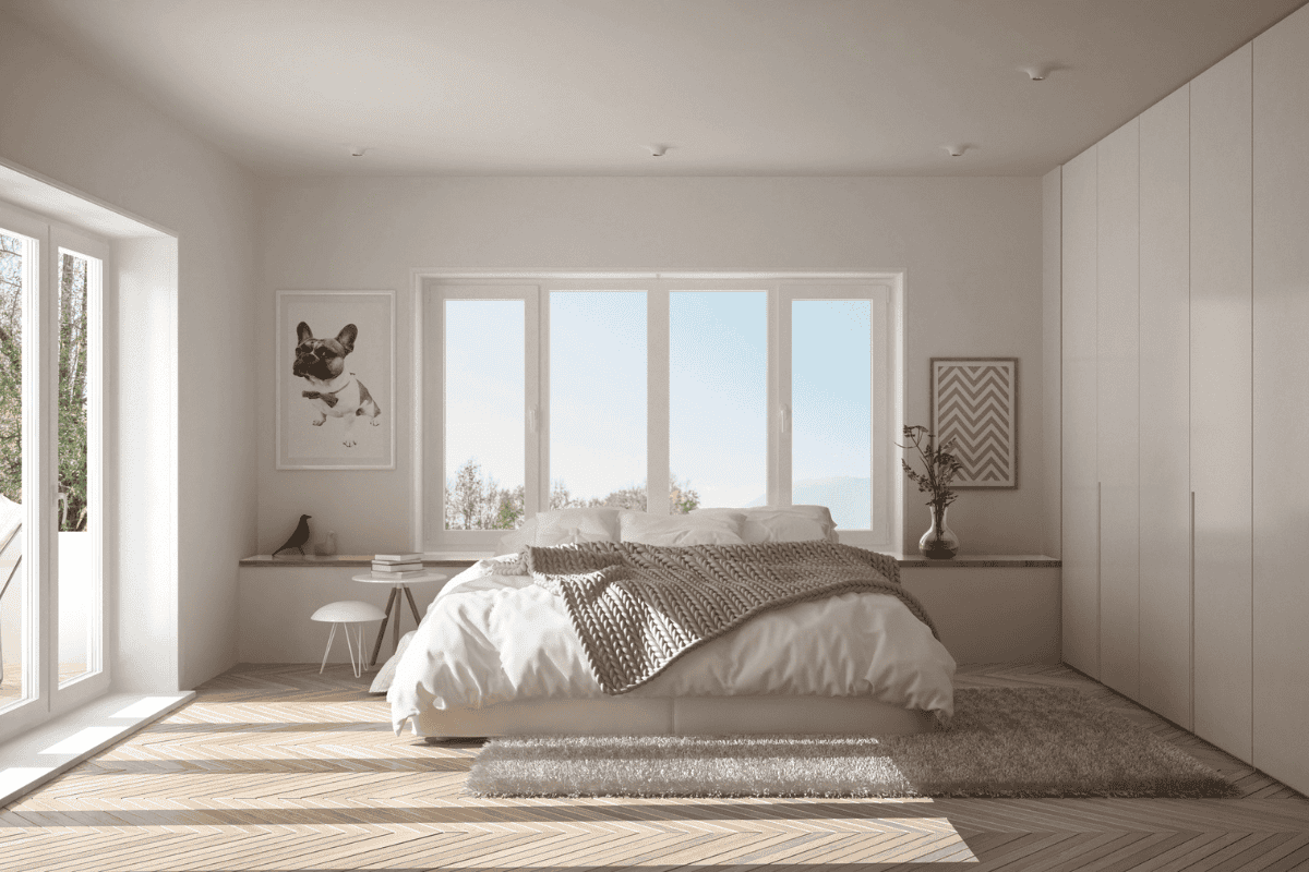 Bright minimalist bedroom with a white bed, built-in wardrobes, light wood floors, and large windows letting in natural light.