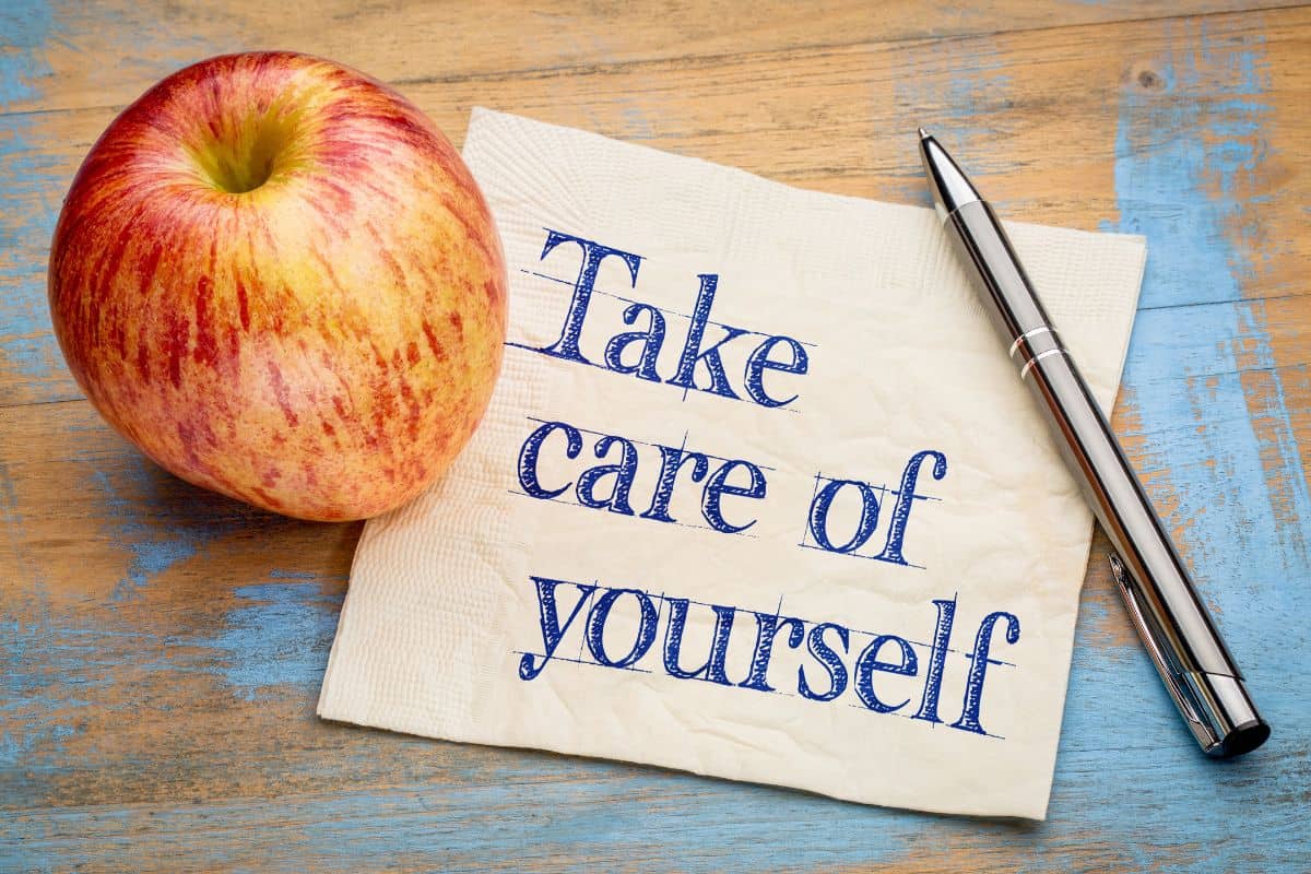 An apple and a pen rest on a wooden table beside a napkin that reads “Take care of yourself.”
