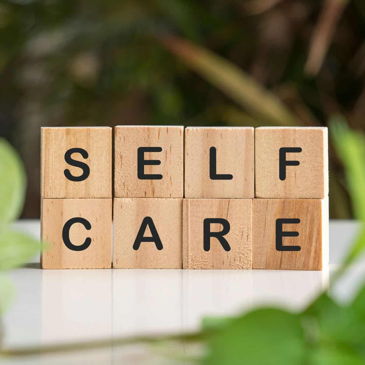 Wooden letter blocks spelling “self care” on a table surrounded by green plants.