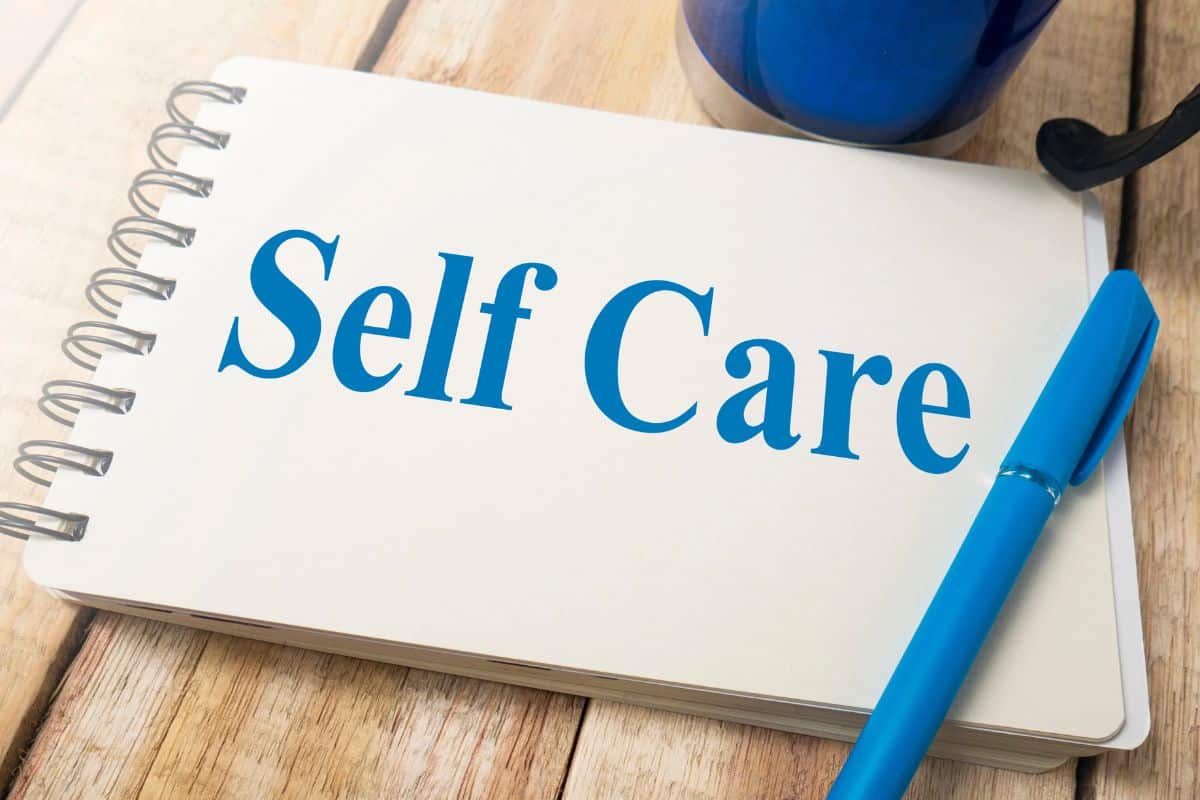 A spiral notebook on a wooden desk displays the words “Self Care” beside a blue pen and mug.