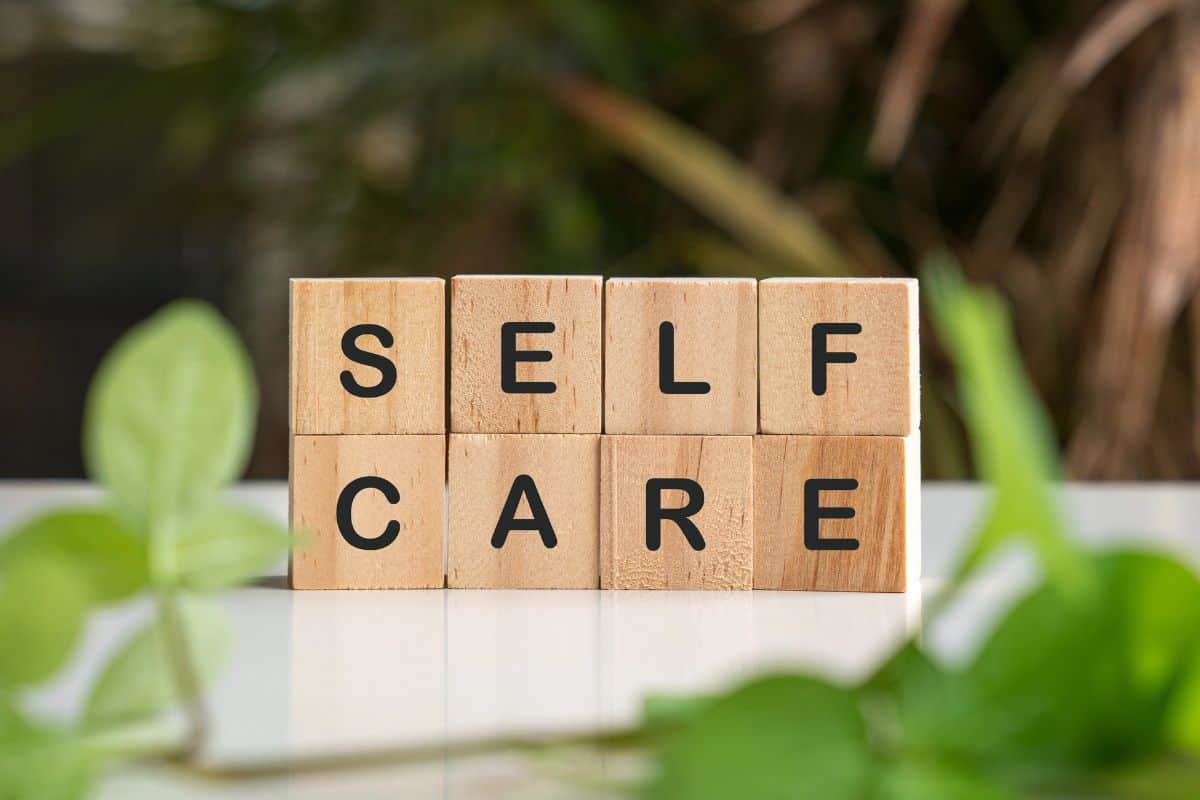 Wooden alphabet blocks spell “self care” on a tabletop, framed by lush green plants.