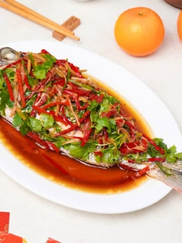 Chinese steamed whole bass topped with ginger, scallions, red chili, and cilantro in soy sauce on a white platter.