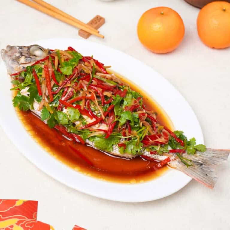 Chinese steamed whole bass topped with ginger, scallions, red chili, and cilantro in soy sauce on a white platter.