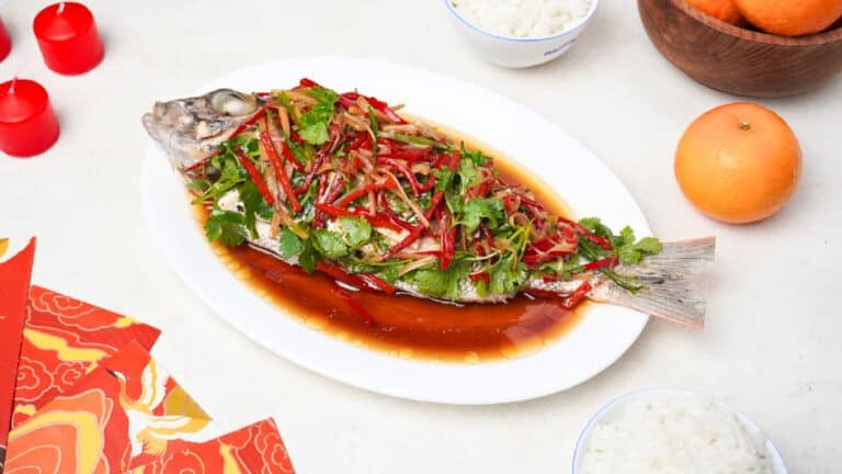 Delicious Chinese steamed whole bass topped with ginger, scallions, red chili, and cilantro in soy sauce on a white platter.