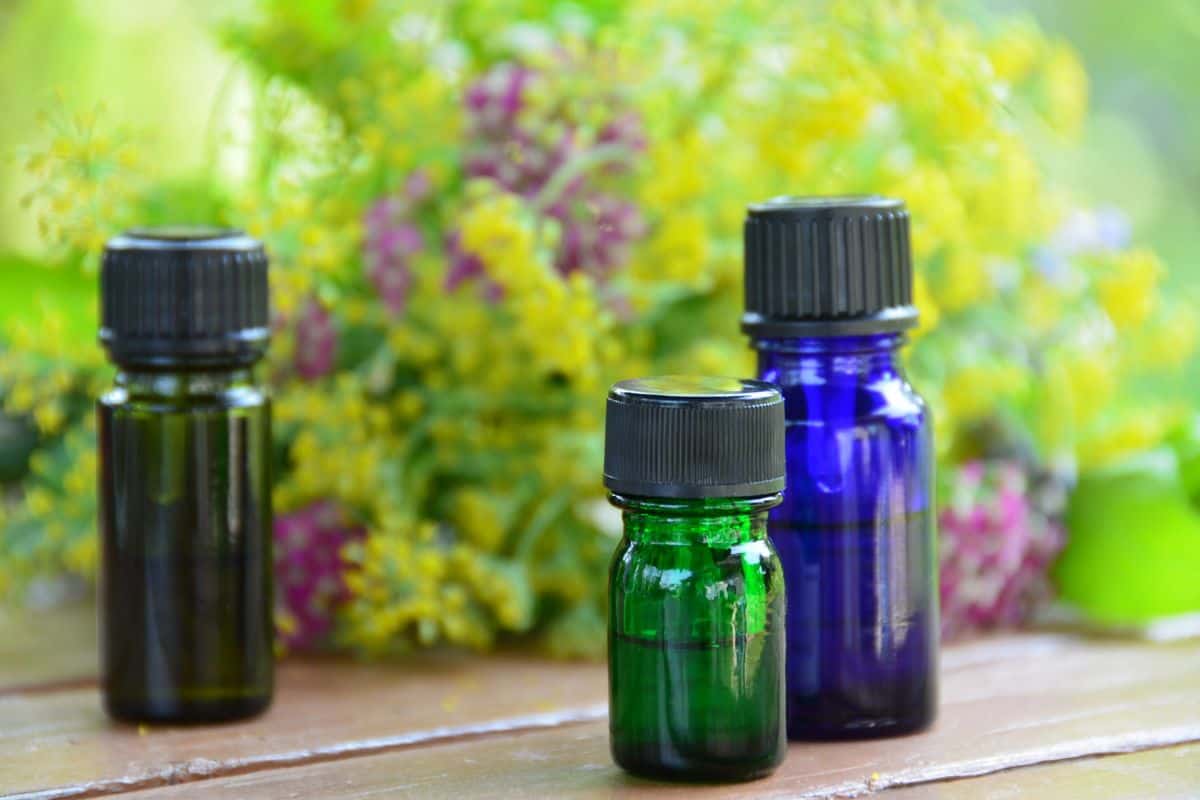 Three small glass bottles of essential oils in green and blue sit on a wooden surface with soft-focus flowers in the background.