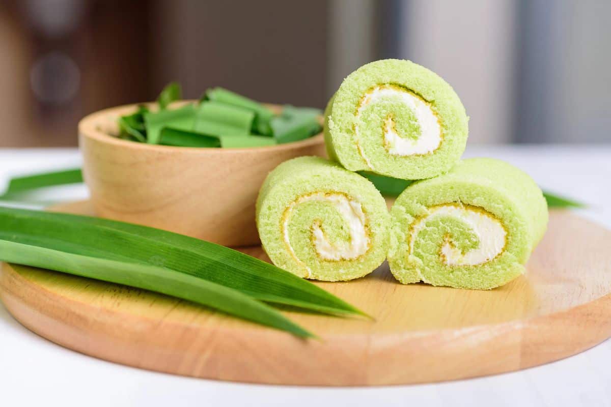 Green pandan Swiss roll cake slices with cream filling displayed on a wooden board with fresh pandan leaves.
