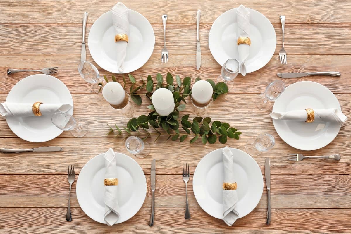 Overhead view of a minimalist table setting with white plates, folded napkins, candles, and greenery centerpiece.