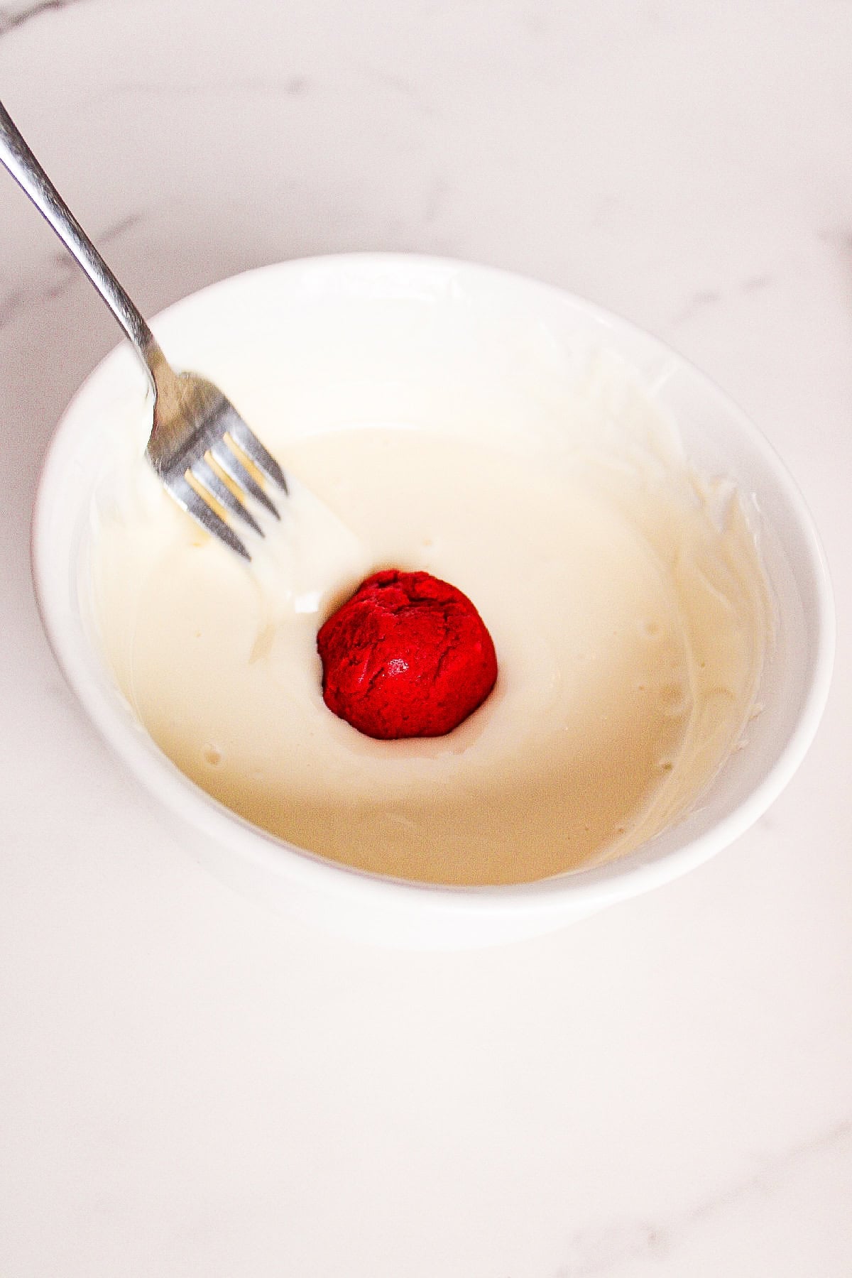 Red velvet cake ball being dipped into melted white chocolate.