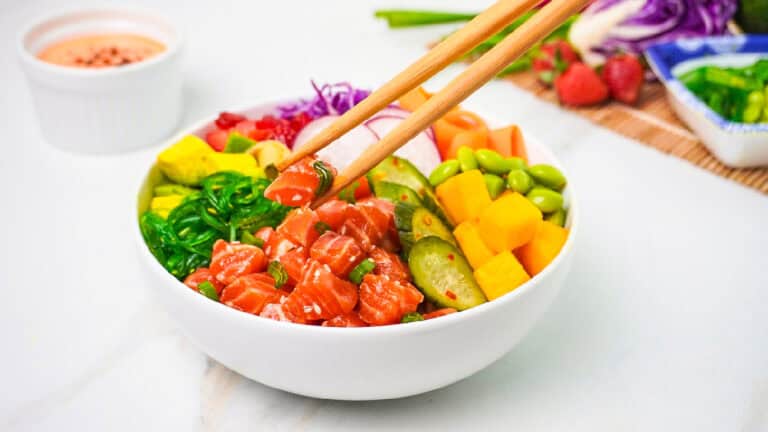 A vibrant salmon poke bowl topped with fresh vegetables and fruit as chopsticks pick up a piece of seasoned salmon.