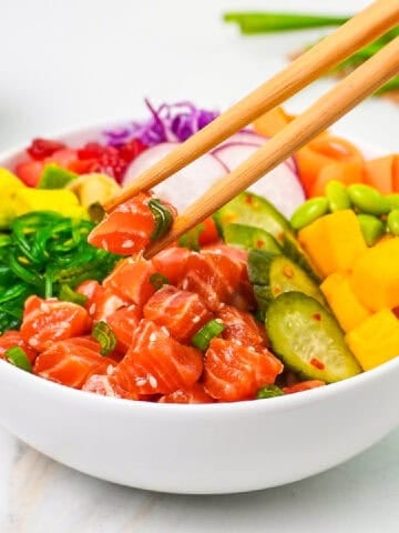 Chopsticks lift marinated salmon from a colorful salmon poke bowl with avocado, mango, cucumbers, edamame, seaweed salad, radishes, and cabbage.