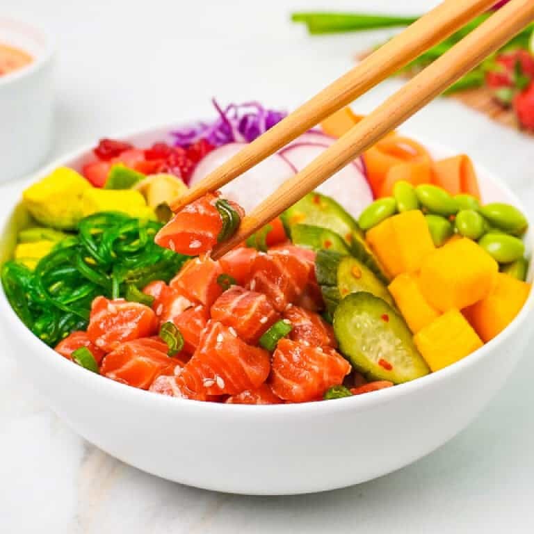 Chopsticks lift marinated salmon from a colorful salmon poke bowl with avocado, mango, cucumbers, edamame, seaweed salad, radishes, and cabbage.