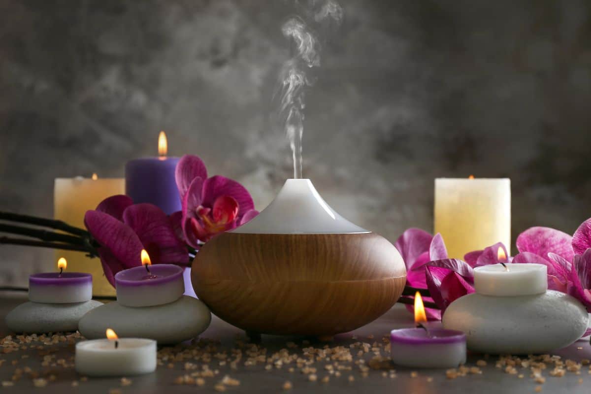 An essential oil diffuser releasing mist surrounded by lit candles, orchids, and spa stones in a calming setting.