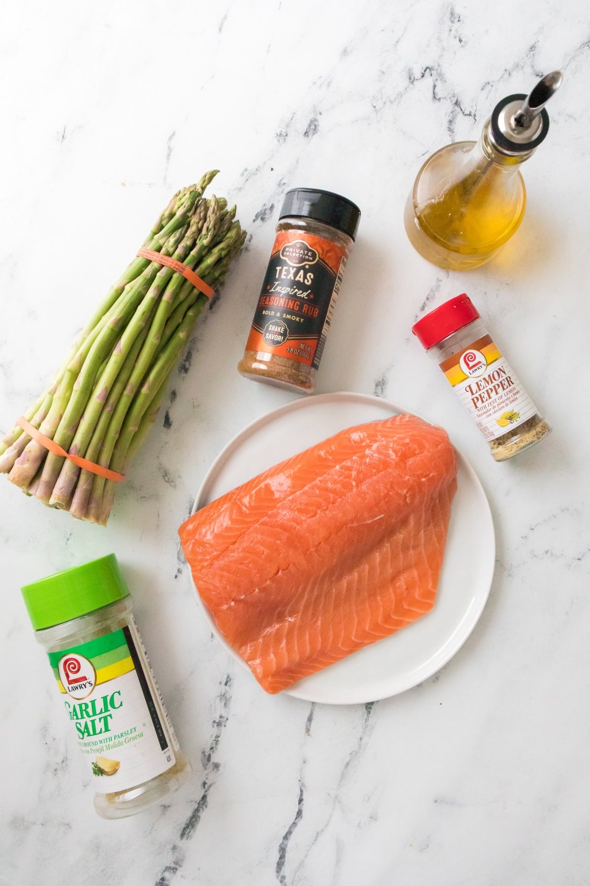Raw salmon fillet on a plate with asparagus, olive oil, garlic salt, lemon pepper, and seasoning for air fryer salmon and asparagus recipe.