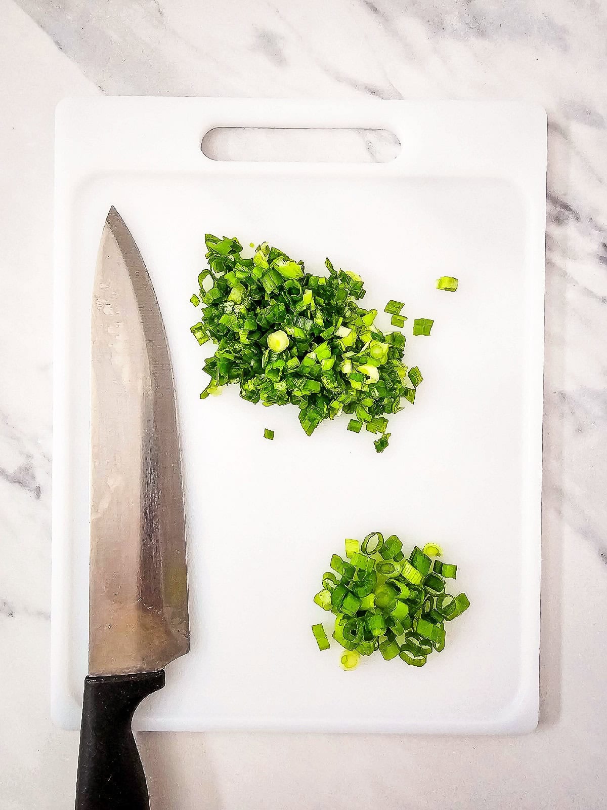 Finely chopped green onions on a white cutting board with a chef’s knife.