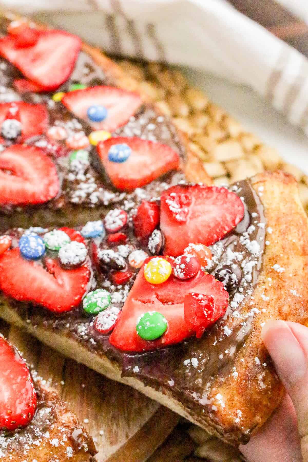 Hand lifting a slice of Nutella dessert pizza topped with strawberries and candy pieces.
